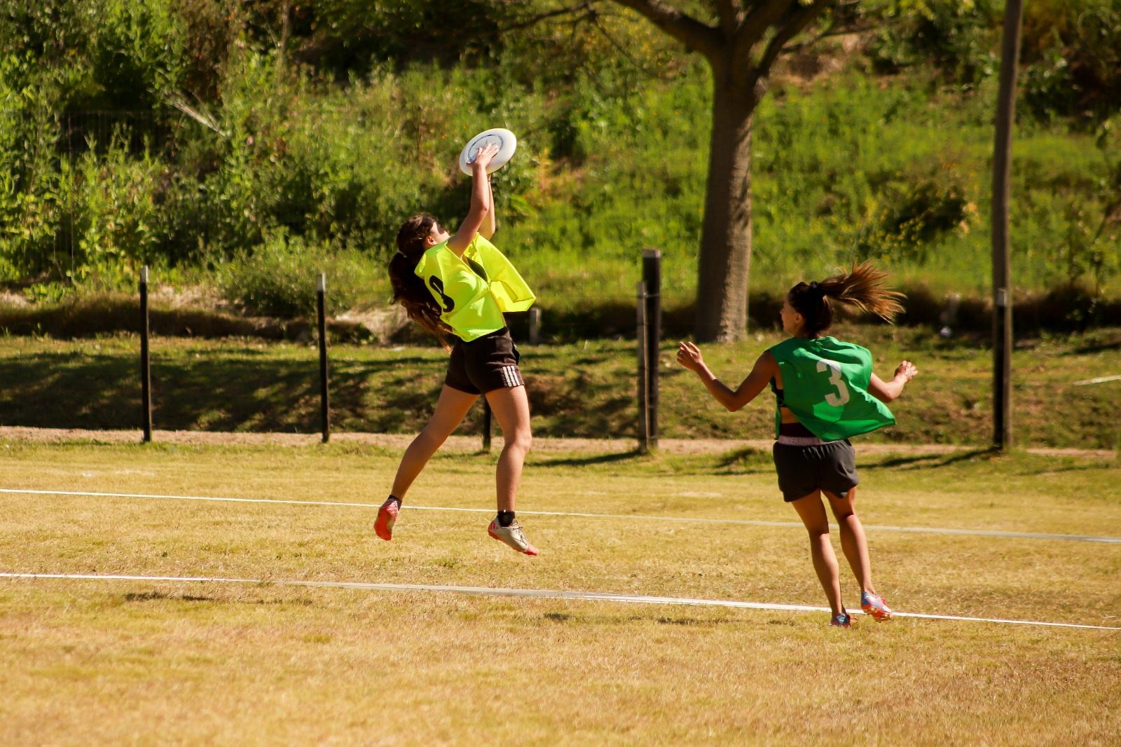 Jugadora con pechera amarilla saltando para atrapar un disco, marcada por una jugadora de pechera verde